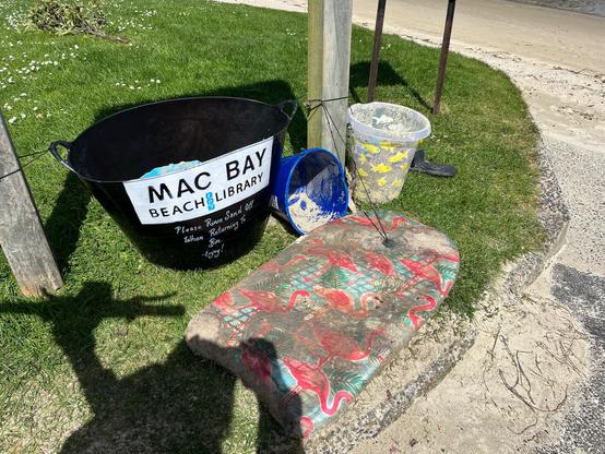 A large black plastic bucket tied to the wooden poles of a sign on some grass, just above a small beach. The box is labelled “Mac Bay Beach Library. Please rinse sand off before returning to bin. Enjoy!” There are some large buckets and a body board next to the bin and inside it (only just visible) some smaller beach toys. It’s sunny and my shadow is visible.