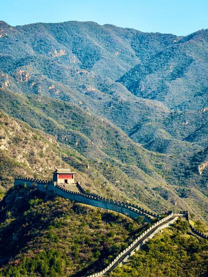 A scenic view of the Great Wall of China winding over the green mountains under a clear blue sky. The sunlight highlights the ancient stone structure and the watchtower perched on the ridge, surrounded by vast, rugged hills fading into the distance.