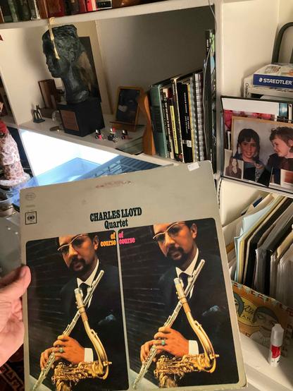 This photo reveals a Charles Lloyd album from an impressive collection [oh please], shown in a personal, lived-in creative space.

The Album: "Of Course, Of Course" by the Charles Lloyd Quartet - featuring a striking cover design with a split image showing Charles Lloyd in formal attire (white shirt, bow tie, and dark suit) holding his flute. The dual image creates a mirrored effect, with colorful "of course" text in orange and pink between the two portraits. Lloyd wears sunglasses and is photographed against a dark background, giving the cover a sophisticated, cool jazz aesthetic. The album is on the Columbia label.

The Setting: This is a creative person's [ha ha] workspace/living area:

° White shelving units filled with books, art supplies, and personal items
° A dark sculptural bust visible on the upper shelf
° Various art books and a Staedtler (art supply) box
° Personal photographs displayed among the collections
° More vinyl records stored in crates/boxes
° The leopard-print decorative item visible again
° Art materials and tools scattered throughout
° A casual, working artist's [???] environment rather than a pristine display space

The View: My hand holds the album, giving scale and a personal touch. The record collection visible in the background suggests this Charles Lloyd album is part of a broader music library, housed within a home workspace used for both listening and creating. [well, sort of]

ALT by Claude, lightly edited and annotated