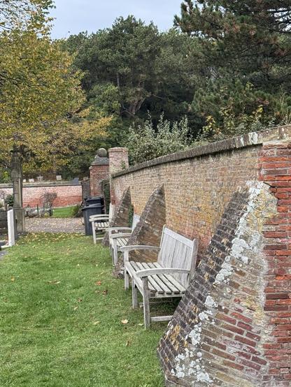 A row of wooden benches is positioned against a brick wall in a park setting, surrounded by green grass and trees. The wall features sections that are angled, covered in moss and lichen. Visible in the background are scattered gravestones and a trash bin

Eine Reihe von Holzbänken befindet sich an einer Backsteinmauer in einer Parkumgebung, umgeben von grünem Gras und Bäumen. Die Wand verfügt über abgewinkelte Abschnitte, die mit Moos und Flechten bedeckt sind. Sichtbar im Hintergrund sind verstreute Grabsteine und ein Mülleimer