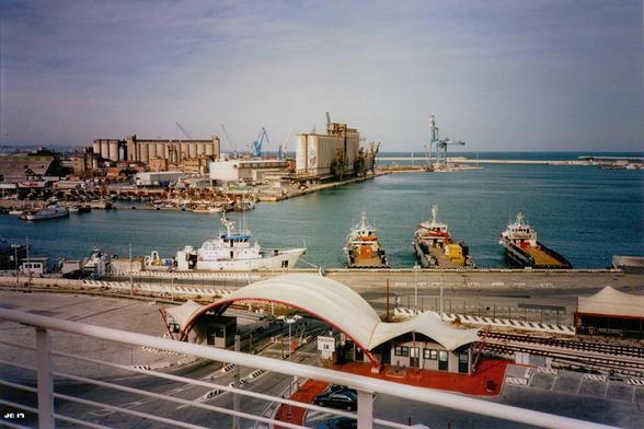 Farbfoto mit Blick über den Hafen von Ancona. Im Vordergrund ein Geländer, darunter Hafengebäude und anschließend Schiffe vor Anker, ein Ausschnitt Wasser, dann weitere Hafengebäude, darunter auch Silos. Über allem ein leicht bewölkter Himmel. Olympus XA auf Kodak Ektar.