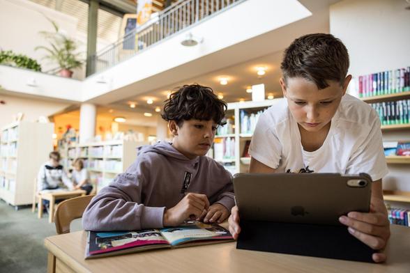 Zwei Jungs sitzen an einem Tisch und schauen gemeinsam in ein Tablet. Auf dem Tisch liegt ein Buch. Im Hintergrund sieht man Bücherregale.