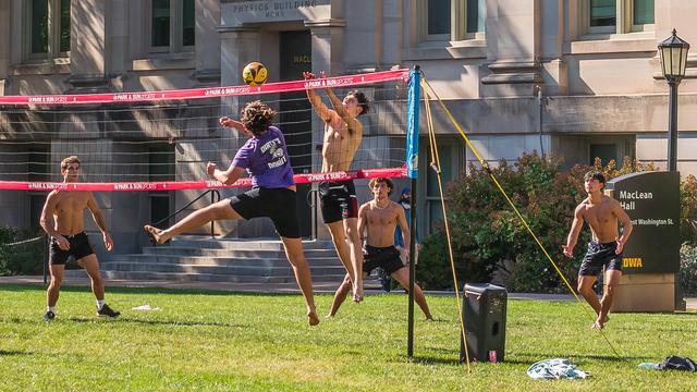 Photograph of a group of young men playing volleyball on a section of the University of Iowa's Pentacrest near MacLean Hall in Iowa City, Iowa. The players have set up a volleyball net on a green lawn in front of MacLean Hall which was the old physics building but is now the mathematics and computer science building. There are five players in the frame with one player on the near side of the net and four players on the far side. The three remaining players on the near side are out of the frame on the left. The player on the near side is in mid air next to the net and has just attempted to spike the ball across the net. Directly across from that player on the far side of another player who is in mid air next to the net has just successfully blocked the opposing player. The three other players on the far side are crouched in a semicircle surrounding the their team member at the the net.