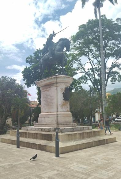 A bronze equestrian statue of Simon Bolivar, a venezuelan national hero, stands on a tall, stepped marble pedestal in a paved plaza with a geometric pattern. Lush trees and a tall palm tree frame the background under a sky with clouds. A pigeon is perched on the pavement in the lower left corner.