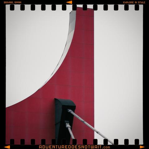 A bold, curved red structure in Bilbao, Spain, framed by modern urban architecture. The striking geometric lines and vibrant color inspire organic cotton adventure apparel with contemporary designs.