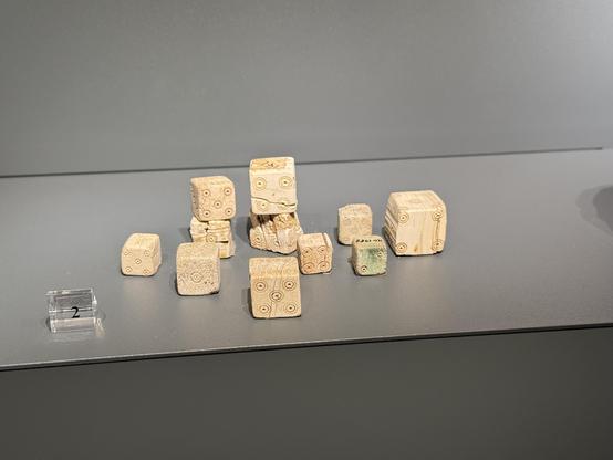 Mehrere etruskische Knochenwürfel in einer Vitrine im Museum. Einige liegen einzeln, einige sind gestapelt. Sie zeigen verschiedene Augenzahlen.