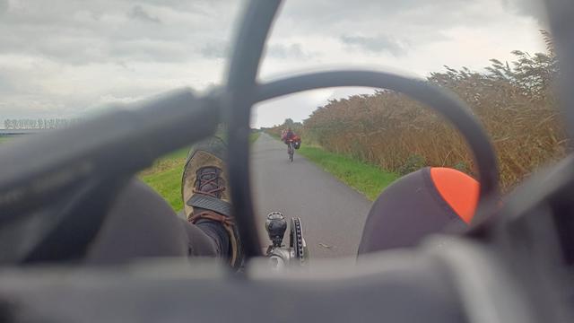 Op een fietspad onder een grijze hemel rijdt een ligfietser voor me. Ze is zichtbaar tussen, heel dichtbij en daardoor uit focus, de remkabels op mijn stuurtje. Mijn beide knieen, mijn linkervoet, voorlicht en kettingblad bevinden zich vooraan in het beeld
