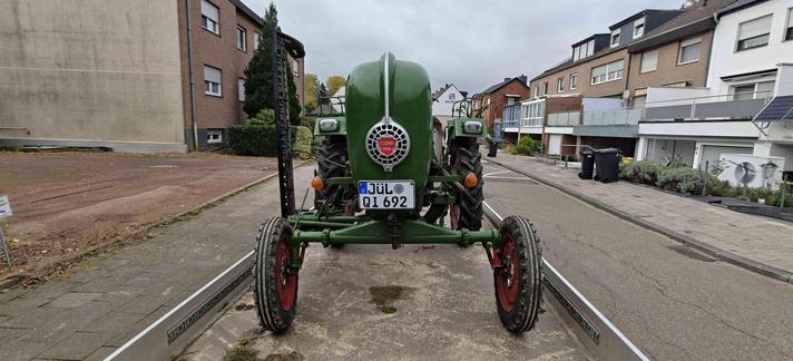 Ein grüner alter Traktor von vorne. Links ist ein Mähbalken (hochkant) sichtbar.
