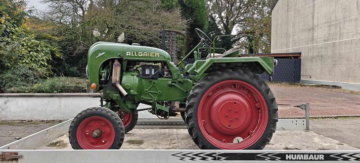 Alter grüner Traktor von links. Man sieht den Motor (links, beim Auspuff), die Batterie (Mitte), Lenkrad und den Fahrersitz (rechts)