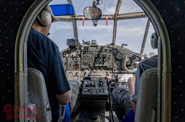 Farbfotografie im Format 3:2 Landscape.
Blick in das Cockpit einer Antonov AN2 und weiter, durch die Scheiben in den blauen, mit wenigen Schleierwolken bedeckten Himmel. Im Cockpit sind viele Knöpfe, Schalter und Hebel zu sehen. Mit dem Rücken zur Kamera und größten teils durch die Trennwand zum Cockpit abgedeckt, links der Pilot mit Baskape und Kopfhörern und rechts der Copilot, ebenfalls mit Kopfhörer.