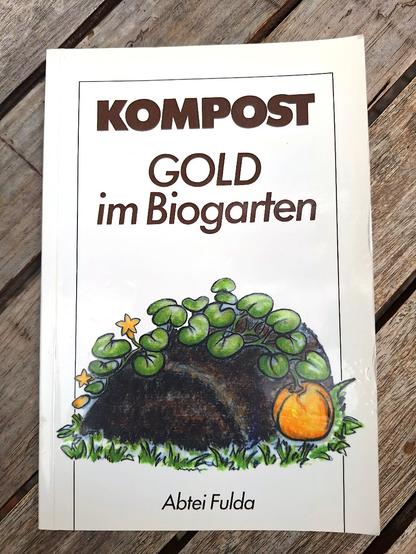 Das Cover einer Broschüre mit dem Text : Kompost - GOLD im Biogarten - Abtei Fulda. In der Mitte eine Grafik von einem Komposthaufen. Die Broschüre liegt auf einem Holztisch mit diagonal verlaufenden Holzleisten.