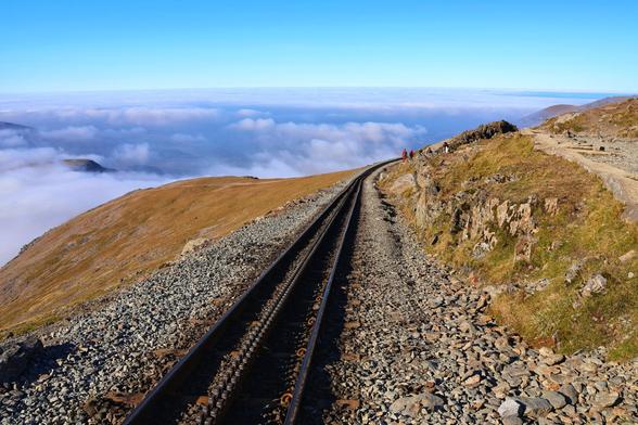 An aerial view of a mountain railway track stretching into the distance. The railway is constructed on a steep, rocky mountainside, with gravel and stones reinforcing the path. The track curves gently as it ascends, disappearing into the horizon. On either side of the track, the landscape is rugged and barren, with patches of grass and shrubs clinging to the rocky terrain.

In the background, a vast expanse of clouds blankets the valleys below, creating a dramatic contrast with the clear blue sky above. The scene is serene and expansive, with a sense of solitude and adventure. A few small figures can be seen walking along the track, adding a sense of scale to the vastness of the landscape.