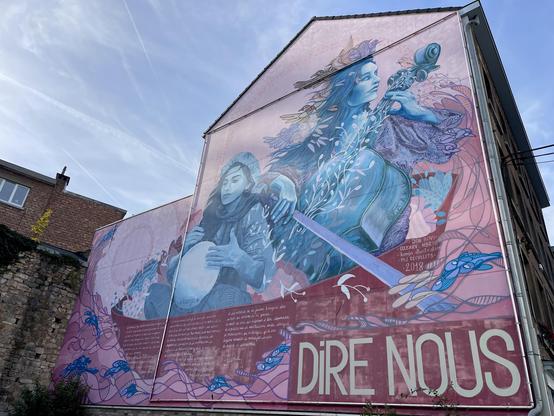 Peinture murale sur une façade latérale de maison. On y voit 2 femmes, l’une jouant d’un violoncelle, l’autre d’un djembé. Un texte parlant de partage de cultures est écrit en dessous de la peinture, mais peu lisible sur la photo, et écrit en grand sur la droite il y a “DIRE NOUS”.