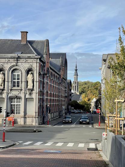 Un carrefour en ville, sur la gauche le coin d’un bâtiment en pierres et briques rouge, avec des statues blanches sur la façade (Palais de Justice). Plus loin dans la rue en face du carrefour on aperçoit un clocher et de nombreux arbres. Le ciel est bleu avec de légers nuages.