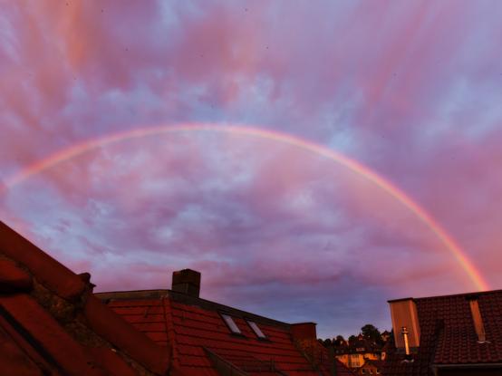 a huge rainbow spanning over dark orange tiled rooftops with chimneys and windows. the sky in the background is blue with pink clouds.