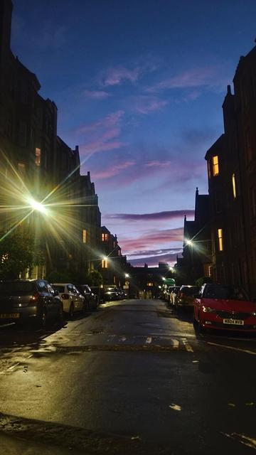 Looking between rows of tall, Victorian tenements on either side of the road as night falls, the dusk sky above a mix of pale blues, purples and pinks