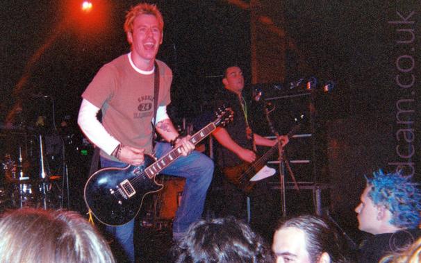 A man with slightly spiky brown hair, wearing a brown t-shirt over a long-sleeved white t-shirt and a blue pair of jeans, playing a black electric guitar attached to a black strap slung over his left shoulder.
He is leaning out over the crowd, his face covered in a massive grin.
The heads of several people can be seen at the bottom of the image, mostly watching the band intently.
In the background, a man wearing a black shirt and black pants can be seen playing a brown and white bass guitar on the right of the frame, next to a rack of black amplifiers, a microphone on a stand in front of him.
A massive drum rig is in the background on the left, under a red stage light.
Black walls at the back of the stage complete the image.