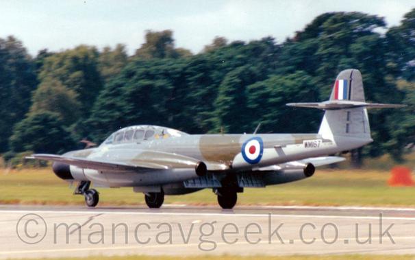Rear view of a twin engined military jet fighter accelerating from right to left along a runway, with small flaps extended at the rear of the wings, between the fuselage and engines..
The plane is covered in a grey and brown camouflage scheme, with pale blue belly, as well as the under-sides of the wings and engine pods.
The wings appear to run through the sides of the engine pods, about a quarter of the way long the wings.
There is a blue, white, and red roundel on the rear fuselage, with the black serial "WM167" further back under the tail.
There is a red, white, and blue fin flash on top of the tail.
A grey runway stretches across the bottom of the frame, with grass on the far side leading up to trees in the distance, under grey sky.