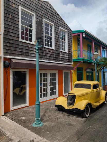 We're in an old city street, narrow and pedestrianised with some lamp posts along the centre and a vintage American car in bright yellow parked up as a display attraction outside a two-storey building with orange lower floor and grey-tiled upper. The building behind is a riot of bright colours - yellow, pinks, greens, blues - and has a colourful balcony on its second floor. It's bright and sunny and feels hot.