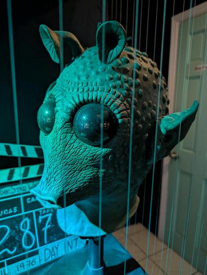 In green-blue lighting we're looking at a movie prop, a prosthetic mask of an alien with a tapering snout for a mouth, two huge eyes, a bobbled skin head with two, short trumpet-like antennae on it, and two pointed ears on the head's side. It is Greedo from the Star Wars films. A clapperboard from a movie is behind.