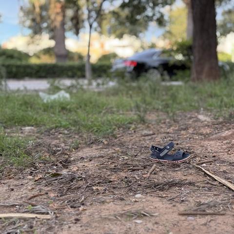 Fotografia d’un parc on es mostra, en primer pla, una única sandalia infantil, enfocada, sobre la terra marró i fulles seques. Cap al fons, l’herba verda es difumina mentre s’allunya de la càmera. Més al fons encara, es ver part d’un carrer, amb la vorera, el tronc d’un gran arbre i un cotxe aparcat a la seua ombra.