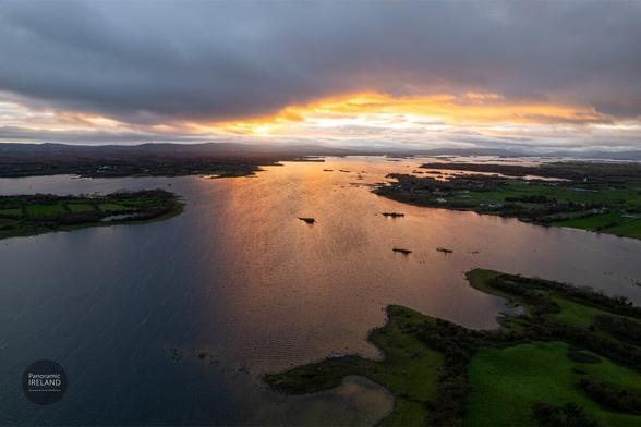 A high-angle, panoramic aerial scene from the west of Ireland in October, capturing a colourful autumn sunset. The view looks over a vast lough (lake) dotted with numerous small, dark islands. A heavy, dark grey sky dominates, but breaks dramatically at the horizon, allowing a brilliant burst of golden, orange, and yellow light to shine through. This sunset creates a bright, shimmering reflection that stretches across the wide, dark blue water. In the foreground and distance, the lough is bordered by rolling green fields and autumn landscapes. By Panoramic Ireland, panoramicireland.com