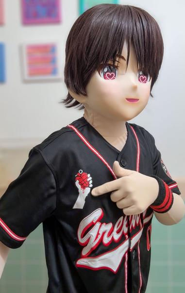 A photo of Billie, my kigurumi OC. He's wearing a baseball jersey themed to Green Day's album American Idiot.

The jersey is mostly black, with white and red trim along the neck and sleeves. On the front, the words Green Day, in a script-y, baseball-y font, is embroidered in white with red borders.

Billie is pointing to a logo on the jersey: the hand holding the heart grenade from the American Idiot album cover.