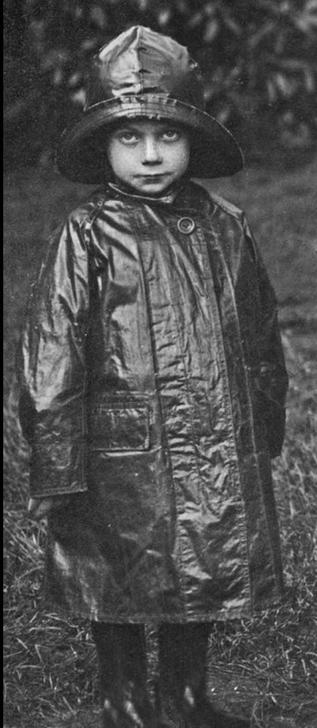 black and white vintage photo of a little kid in a heavy duty raincoat, and wearing a sou’wester rainhat. the front rim of the rain hat is curved up to show the kid’s face, and incidentally creating a rain gutter that would funnel rainwater to the back. the child is looking up at the camera with a fixed, slightly exasperated expression. behind the child, shrubs or trees.  no idea who owns the photo -  when i followed the link i kept ending up at some religious website, and din;t want to hang around. i suspect the person in the photo was born a few years before i was… maybe 1930s
