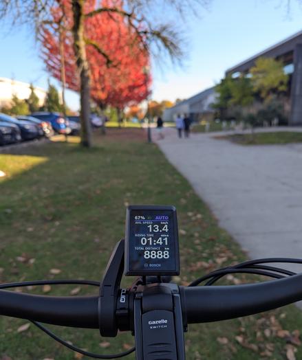 A photo of a bike odometer reading "8888". There's a multiuse path on the right, a parking lot on the left. In the distance are trees with brilliant red and orange colours. The sky is blue overhead.