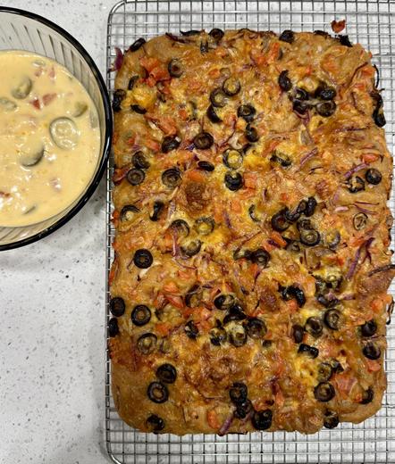 A baked focaccia with olives, onions and tomatoes sprinkled throughout. Next to it is a bowl of cheese sauce with jalapeño slices and tomatoes in it