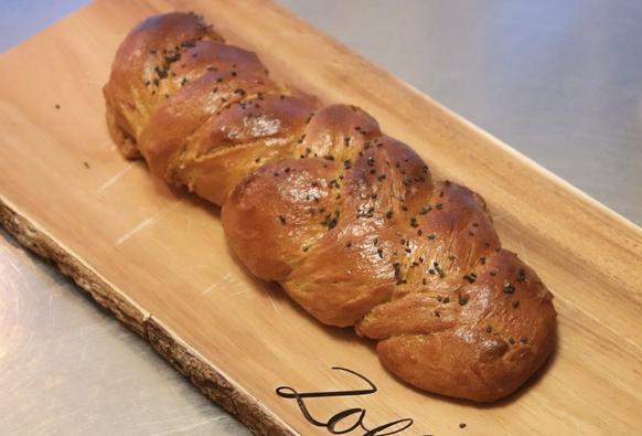 a glazed challah sprinkled with black sesame seeds. the challah is on a wooden cutting board that's on a metallic surface.