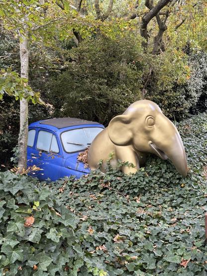 Ein blauer Kleinwagen ist halb im Erdreich versunken. Aus der Motorhaube steigt ein goldener Elefant.