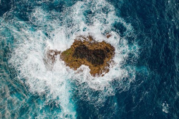 A "from above" drone photo of a small rocky outcropping in the ocean, surrounded by blue water and waves