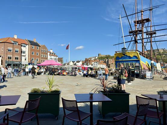 A sunny day in Whitby features a busy harbor square with market stalls selling bags and accessories under pink canopies, groups of people walking and browsing, and a replica sailing ship moored nearby with a colorful blue and yellow stern. The Union Jack flies on a flagpole at the center, with rows of brick and stone buildings along the edge of the square, and Whitby Abbey visible on the hill in the background. In the foreground, tables and chairs are set behind planters, suggesting a café area overlooking the lively scene.