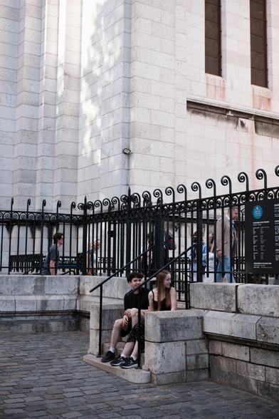 portrait format photo of 2 teens sitting on a small stone stair. A black fence is behind them on top of the 5ish steps stair, white walls of the Sacré Cœur behind the fence. The girl with long dyed hair in dark ginger is turning her head behind her to look at someone, the boy with short fluffy black hair was also looking but caught the camera and is turning his face to it. Behind the fence some tourist are walking to the right of the pic
