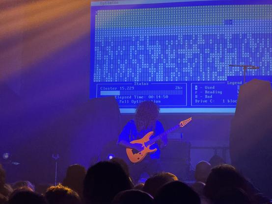 Guitarist from Master Boot Record with big curly hair holding an Ibanez reverse headstock guitar in coral pink, glowing slightly under stage lights. DOS-era output of defrag.exe (I think) projected onto a screen in the background.