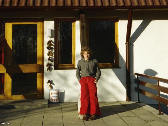 Farbfotos aus den Siebzigerjahren, das einen jungen Mann mit langen Haaren, einem grauen Hemd und einer breiten roten Hose vor einem Gebäude mit Langnese-Papierkorb neben dem Eingang links zeigt.