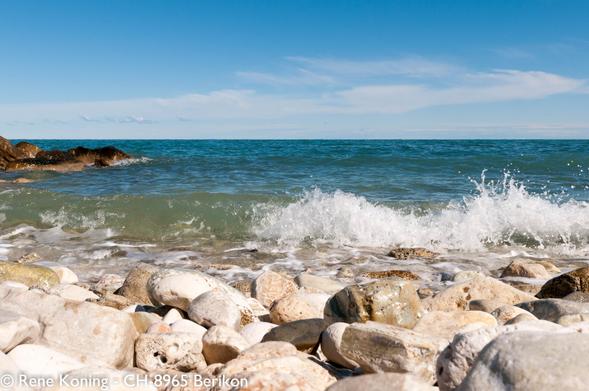 Eine kleine Welle rollt an der Adriaküste an einem Felsenstrand. Schaumkrone zerbricht. Links im Hintergrund ragt eine klein Landzunge in das Wasser. Der Himmel ist Hellblau mit einzelne Wolken