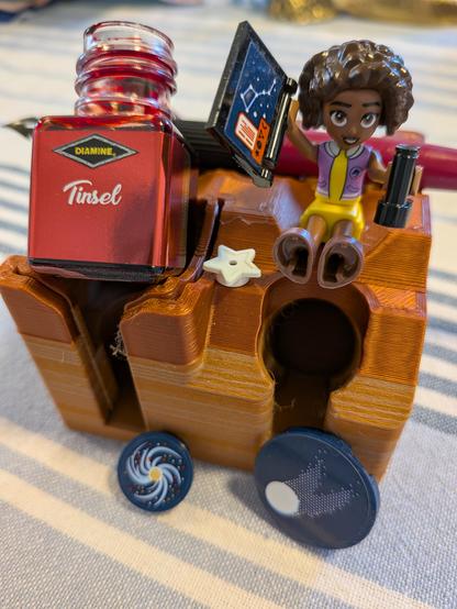 A bottle of Diamine Tinsel ink sits on a stand, and beside it sits a Lego minifriend Aliya with a star chart showing Orion. Some Lego accessories of a star, comet and Galaxy are also present, and Aliya is holding binoculars.