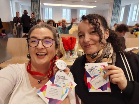 KI-generiert über @altbot, menschlich überarbeitet: Zwei Personen stehen in einer Menschenmenge. Beide tragen Namensschilder, wobei eine Person ein Namensschild mit dem Text "Roxanne Vierhaus" und die andere mit dem Text "Xenia Kitaeva" trägt. Beide Personen tragen rote Umhängebänder. Die Person auf der linken Seite blickt direkt in die Kamera und lächelt. Die Person auf der rechten Seite blickt ebenfalls in die Kamera. Im Hintergrund sind mehrere andere Personen zu sehen. Der Hintergrund ist unscharf. Der Text "ZUGANG GESTALTEN" ist auf beiden Namensschildern zu lesen. Ein digiS-Button befindet sich auf dem Umhängeband auf der linken Seite.