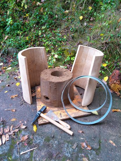 Zwei halbe Holzschalen, die zusammengesetzt eine Eimerform ergeben, stehen um den Lehmofen in der Mitte herum. Die beiden Stahlreifen sind angelehnt, ein Setzhammer liegt davor. Den Hintergrund bildet ein grüner Hang.