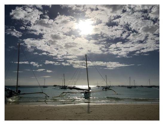 Photo taken on an island beach, looking out to sea. Several small sailboats, with long outriggers extending from their sides, float offshore with their sails furled. The central boat casts the shadow of its mast onto the sand in the foreground. The midafternoon sun at top center frame shines through some clouds and casts glittering light on the waves.