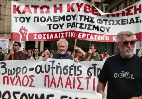 Una manifestazione di protesta con una folla di persone che tengono striscioni. Un grande striscione rosso e bianco in alto recita in greco: "GIÙ IL GOVERNO DELLA GUERRA, DELLA POVERTÀ, DEL RAZZISMO" e "PARTITO SOCIALISTA DEI LAVORATORI", con un simbolo di pugno alzato. Un uomo anziano con i capelli bianchi al centro tiene un altro striscione bianco che recita in greco: "Aumenti di 3 ore a Pylos/Palestina".
