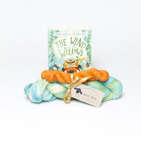 Picture shows a sock set of yarn consisting of a large skein which is a variety of greens and blues and a smaller skein which is orange. They are tied together with gold ribbon. Behnd the yarn, stood on its end is a copy of the book, The Wind In the Willows.