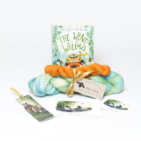 Picture shows a sock set of yarn consisting of a large skein which is a variety of greens and blues and a smaller skein which is orange. They are tied together with gold ribbon. Behnd the yarn, stood on its end is a copy of the book, The Wind In the Willows. In front of the yarn from left to right is a bookmark, a postcard and a sticker.