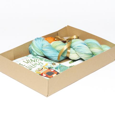 Picture shows a sock set of yarn consisting of a large skein which is a variety of greens and blues and a smaller skein which is orange. They are tied together with gold ribbon. They are inside an open A4 box al;ongside a copy of the bookj, The Wind in the Willows.