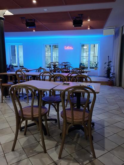 Vista interior de un restaurante o bar con un ambiente moderno y rústico. El espacio está lleno de mesas de madera clara, muchas de ellas cuadradas o rectangulares, y sillas de madera curvada de estilo clásico con asientos de rejilla. El suelo es de baldosas claras con un patrón irregular. Al fondo, una pared blanca presenta tres ventanas grandes con vistas al exterior. Sobre estas ventanas, una distintiva tira de luz de neón azul brillante ilumina horizontalmente la parte superior de la pared. Debajo del neón azul, un cartel de neón rosa escribe la palabra 'Laba.' con un punto. El techo tiene paneles acústicos marrones oscuros.