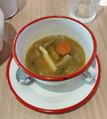 Un tazón de sopa de verduras en un tazón blanco con un borde rojo esmaltado, colocado sobre un platillo a juego. La sopa tiene un caldo espeso de color amarillo verdoso y contiene trozos grandes de verduras, incluyendo una rodaja redonda de zanahoria naranja brillante, un tallo largo de espárrago blanco y otros vegetales pálidos y rectangulares, posiblemente patatas o nabos, junto con pequeños trozos verdes. El tazón y el platillo están sobre una mesa de madera clara. En el fondo, a la derecha, se ve un vaso de cristal texturizado vacío, y a la izquierda, una servilleta blanca o un objeto similar.