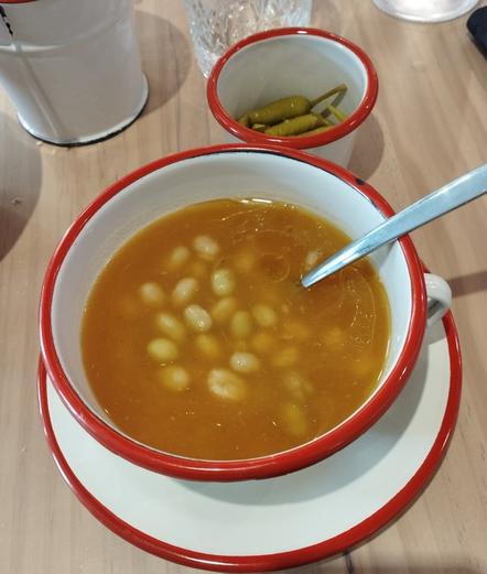 Un cuenco blanco con un borde rojo, lleno de sopa o guiso de alubias blancas en un caldo de color naranja rojizo. Numerosas alubias blancas o de color crema flotan en el caldo, y una cuchara plateada descansa en el cuenco. El cuenco está sobre un plato a juego. A su lado, un pequeño recipiente blanco con borde rojo contiene varias guindillas verdes. En el fondo se aprecian un vaso de cristal texturizado y parte de una jarra blanca. Todo se encuentra sobre una mesa de madera clara.