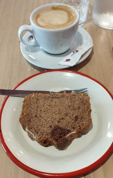 La imagen muestra un primer plano de una mesa de madera clara con un café y un trozo de bizcocho. En la parte superior, una taza blanca de café con leche con espuma y un patrón en espiral, se apoya sobre un platillo blanco. Junto a la taza, hay un sobre de azúcar blanco. En la parte inferior, un plato blanco con un distintivo borde rojo contiene un trozo grande de bizcocho de color marrón oscuro, que parece denso y con posibles trozos de chocolate. Un tenedor plateado descansa sobre el bizcocho.